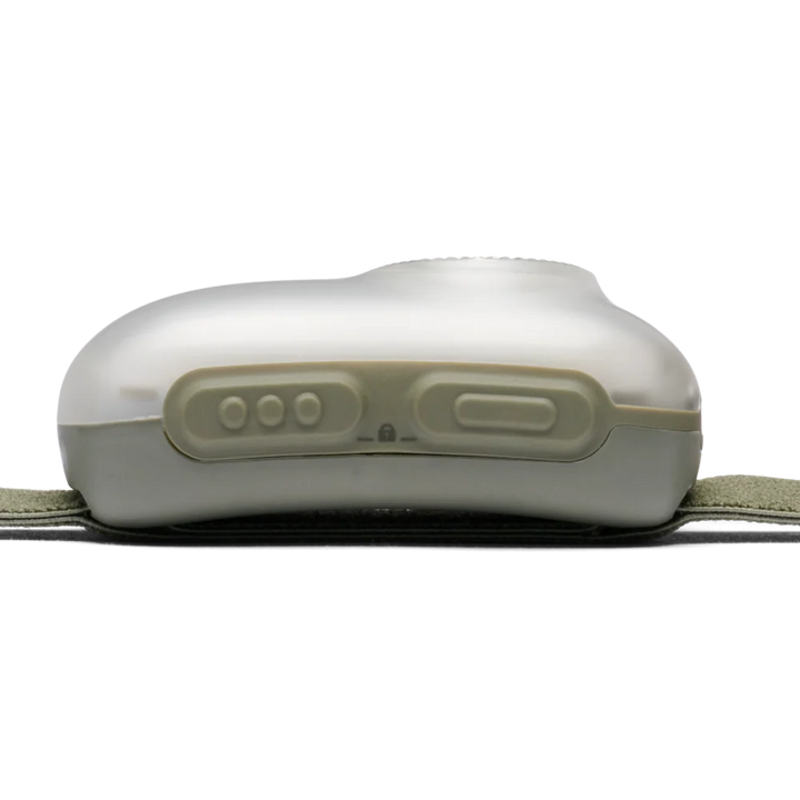 A close-up of a white electronic control unit with three round buttons and one larger rectangular button on a light gray surface, resting on a dark strap with an olive green fabric band visible at the sides.