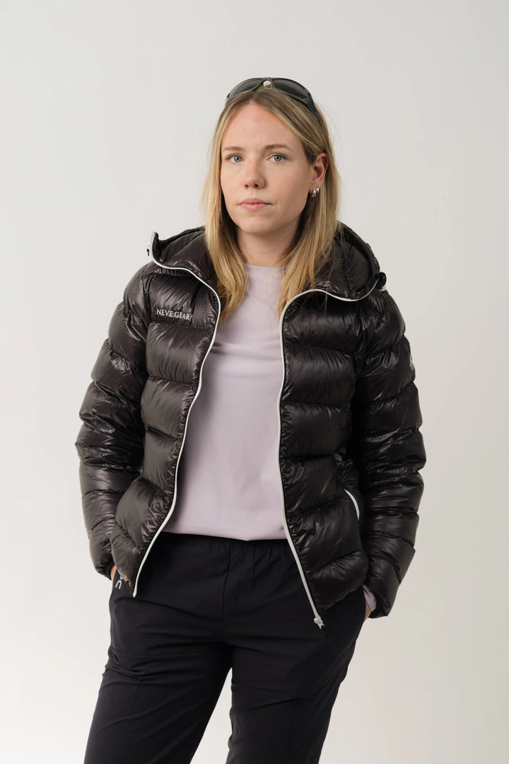 A woman modeling a black hooded puffer jacket with white piping and a small white logo on the left chest, paired with a light pink top and black pants, against a plain light gray background.