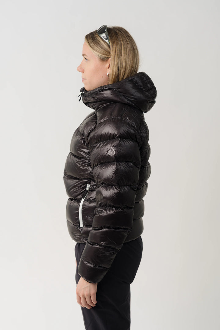 A woman shown in profile wearing a shiny black puffer jacket with a high padded collar and black pants, photographed against a plain light gray studio background.