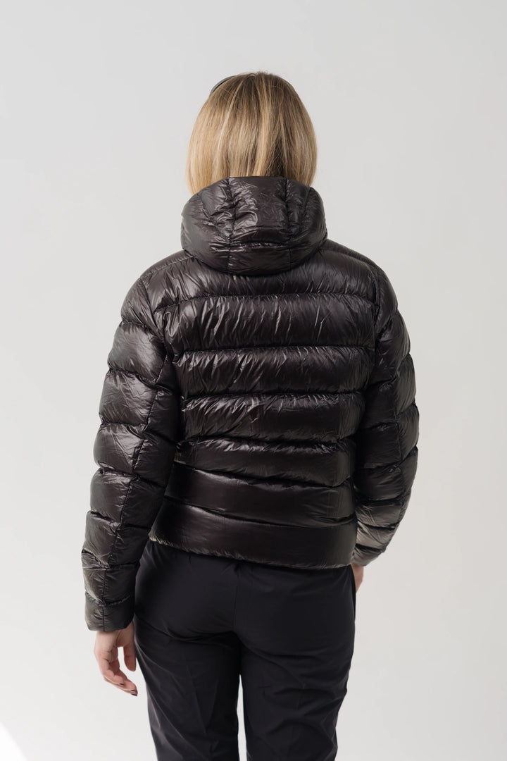 A woman viewed from behind wearing a shiny dark brown puffer jacket with horizontal quilting and a hood, paired with black pants, against a light gray studio background.
