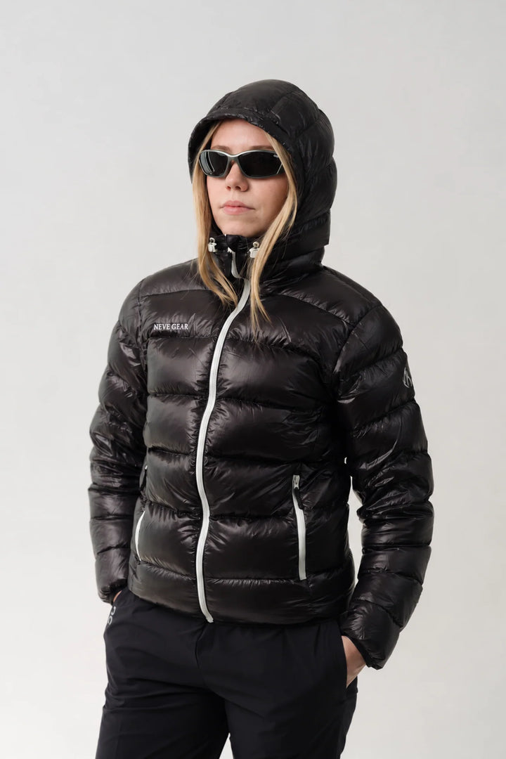 A woman wearing a shiny black hooded puffer jacket with silver zipper accents, black pants, and sunglasses, photographed against a plain light gray studio background.