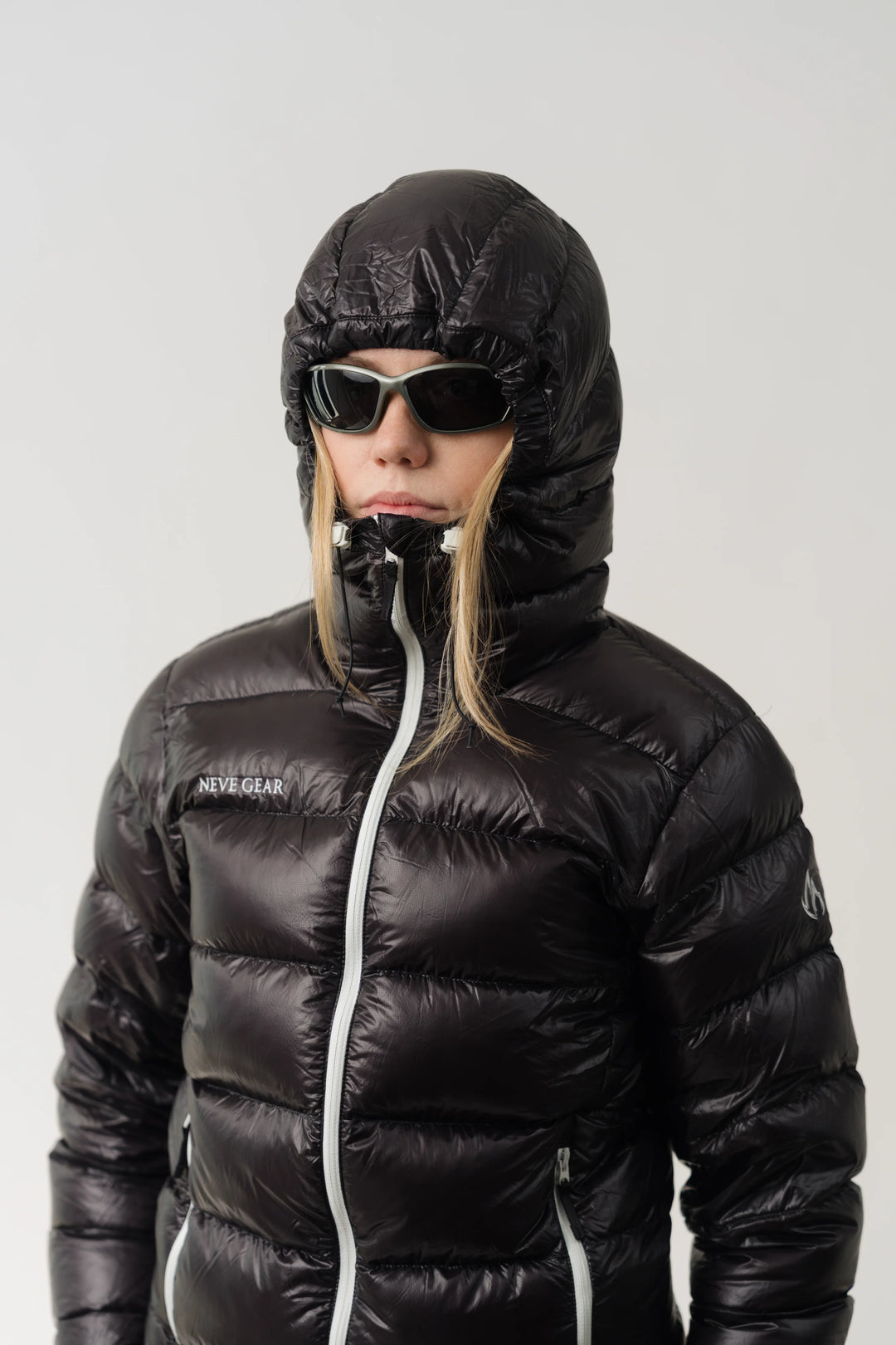 A model wearing a shiny black puffer jacket with a hood, black sunglasses, and a black neck warmer, set against a plain light gray background. The jacket has a white zipper and a small white logo on the left chest that reads 'NEVE GEAR'.