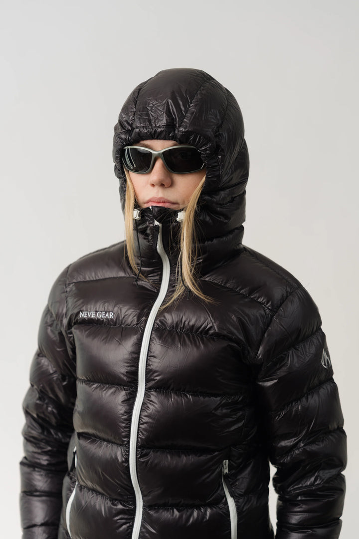 A model wearing a shiny black puffer jacket with a hood, black sunglasses, and a black neck warmer, set against a plain light gray background. The jacket has a white zipper and a small white logo on the left chest that reads &