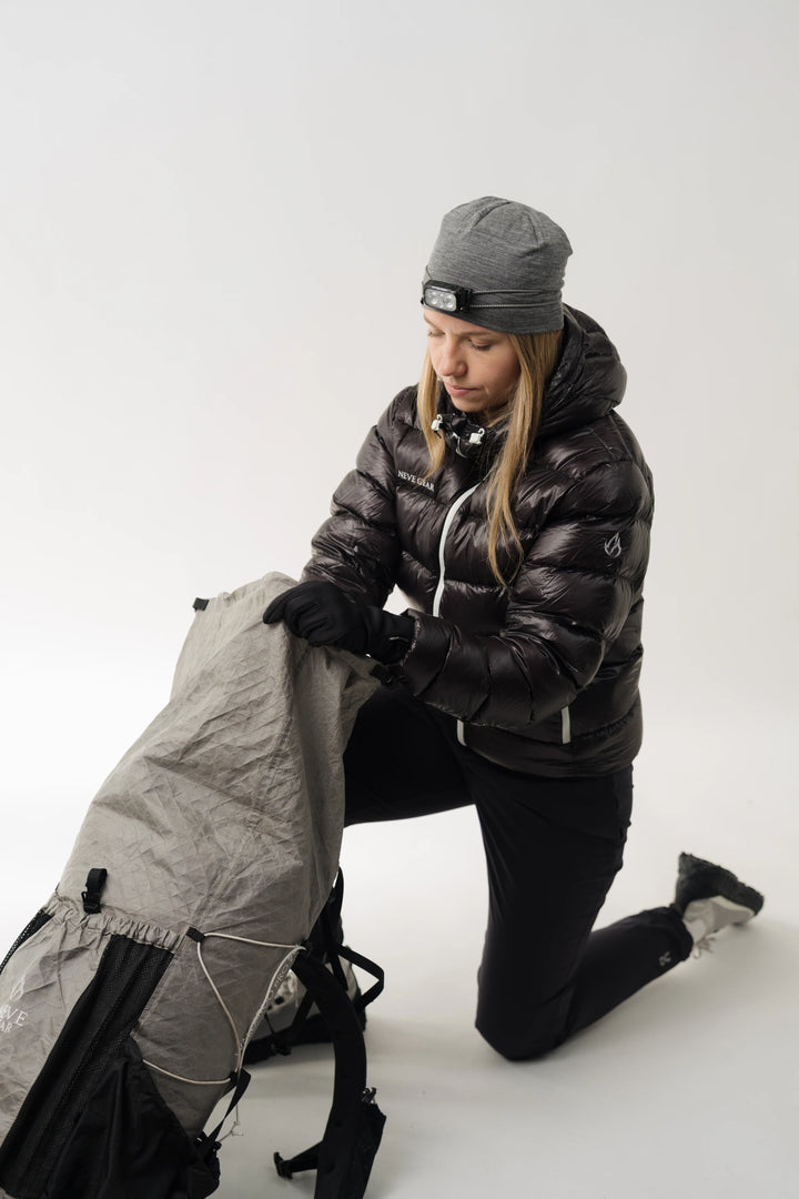 A woman kneeling on a white studio floor wearing a black puffer jacket, black pants, and a gray beanie, while packing a large gray and black backpack.