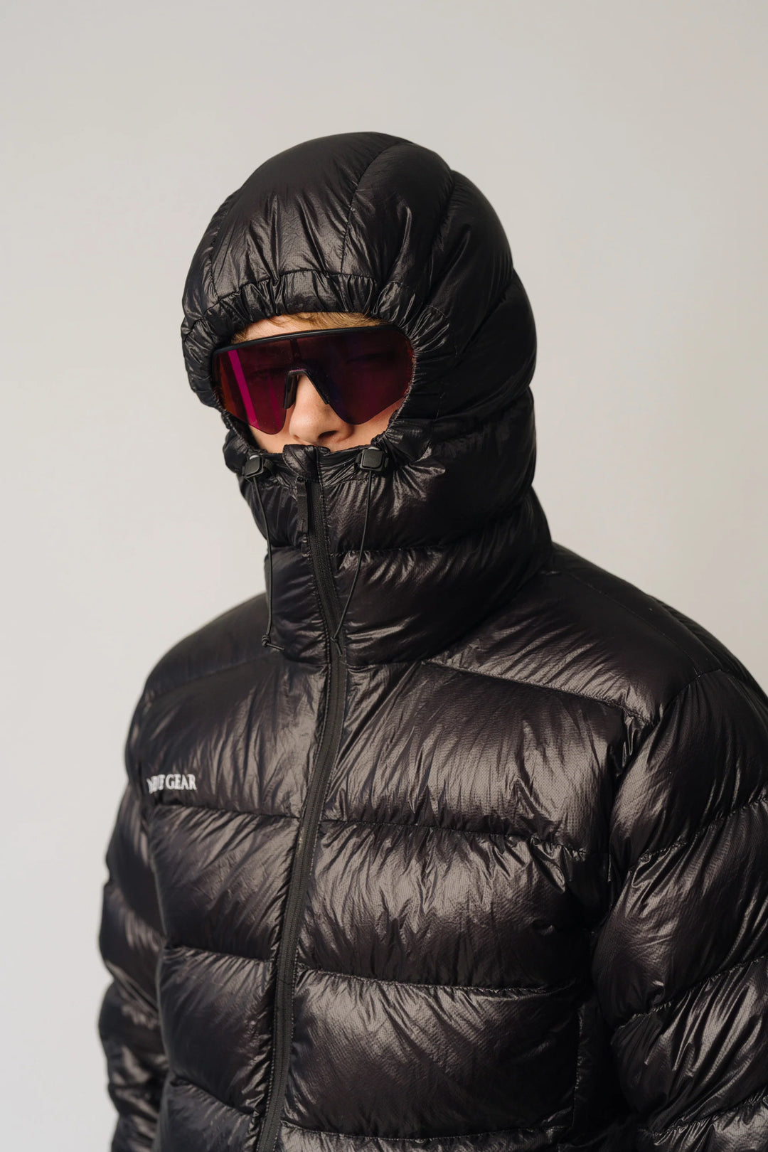 Person wearing a black puffer jacket with a hood and red sunglasses on a plain background