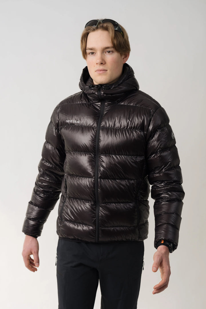 A man wearing a shiny dark black puffer jacket with a high collar and front zipper, paired with black pants, photographed against a light gray studio background.
