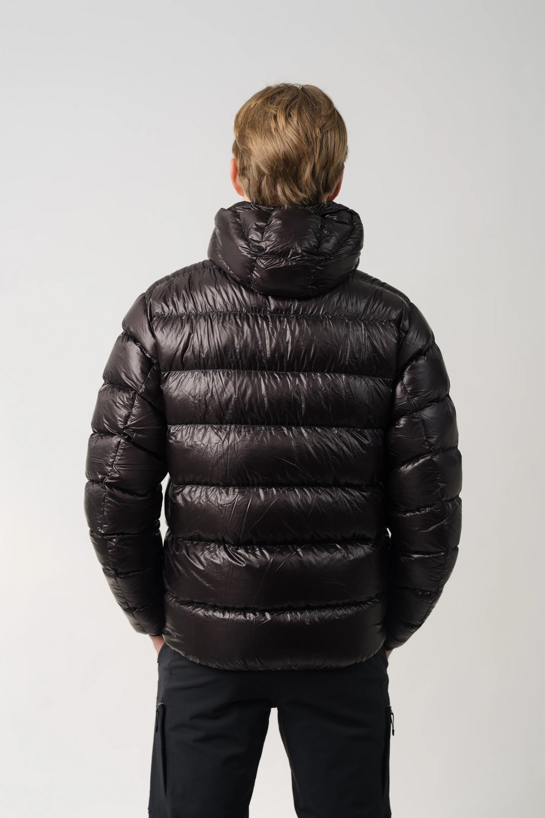 A man viewed from behind wearing a shiny black hooded puffer jacket and black pants against a light gray studio background.