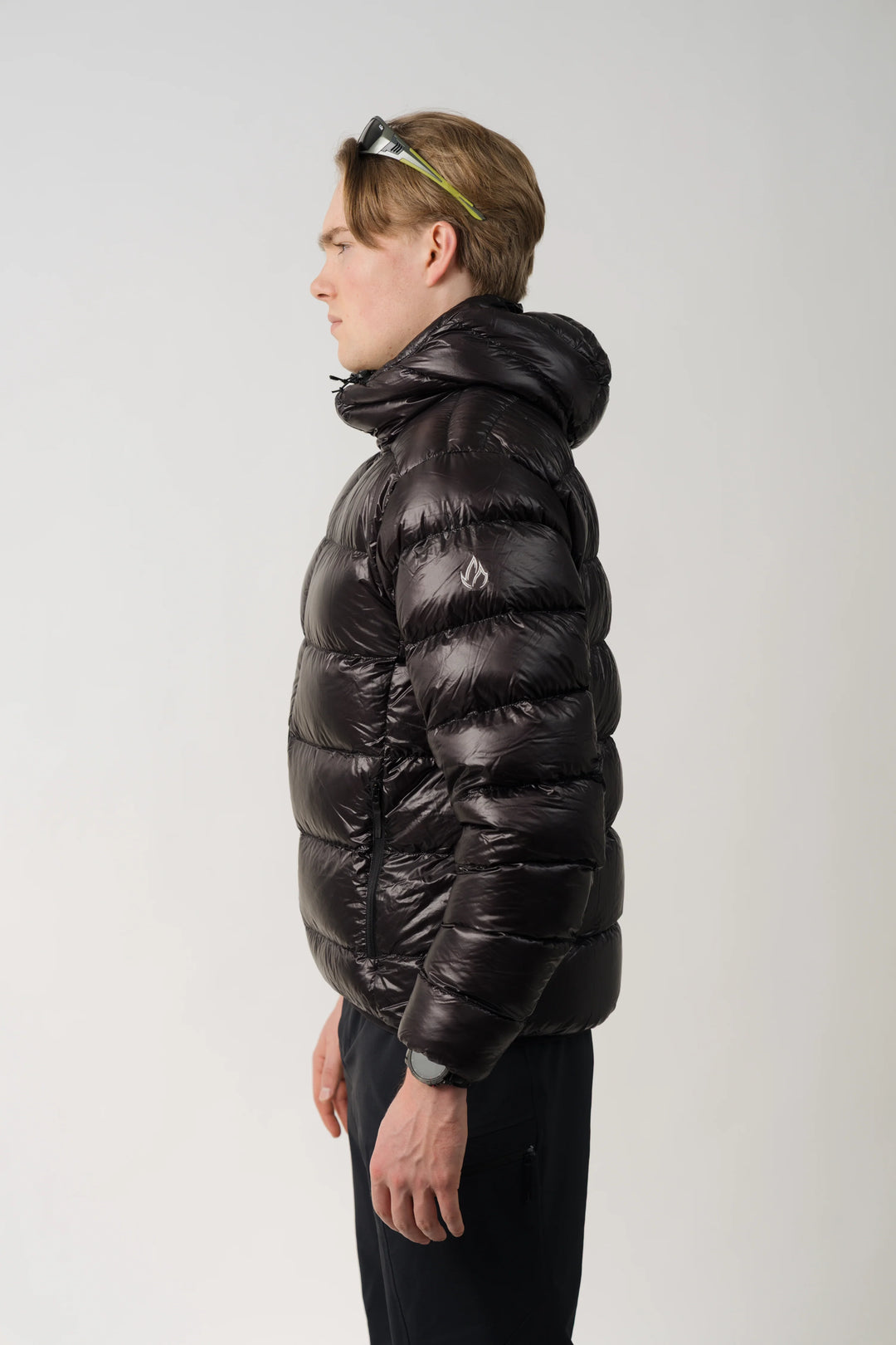 A man in profile wearing a shiny black hooded puffer jacket and dark pants, photographed against a plain light gray studio background.