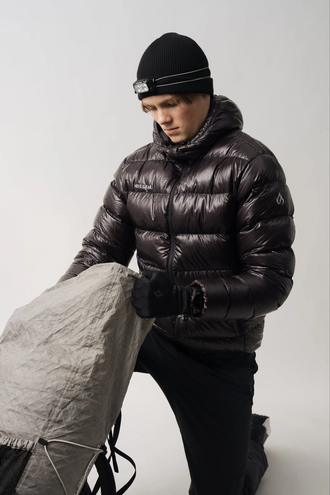 A man wearing a black beanie with a headlamp and a shiny black puffer jacket kneels while holding a gray tent bag against a plain light gray studio background.