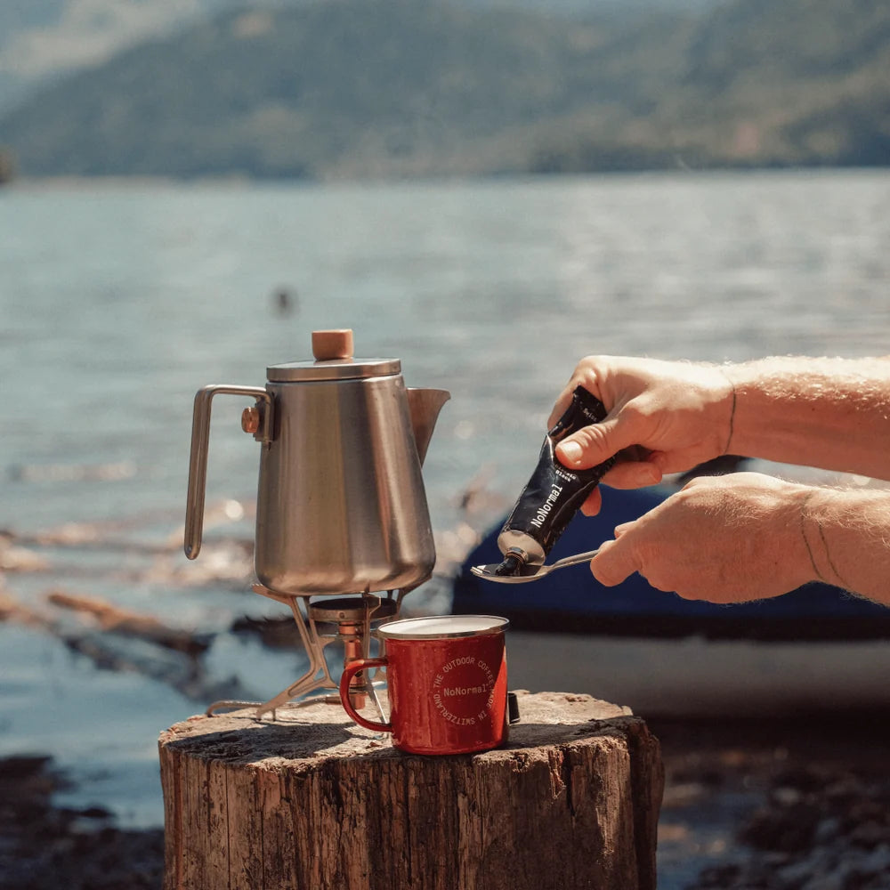 Outdoor setting on a body of water with a kettle boiling on a camping stove and a mug on a wooden stump. A person&