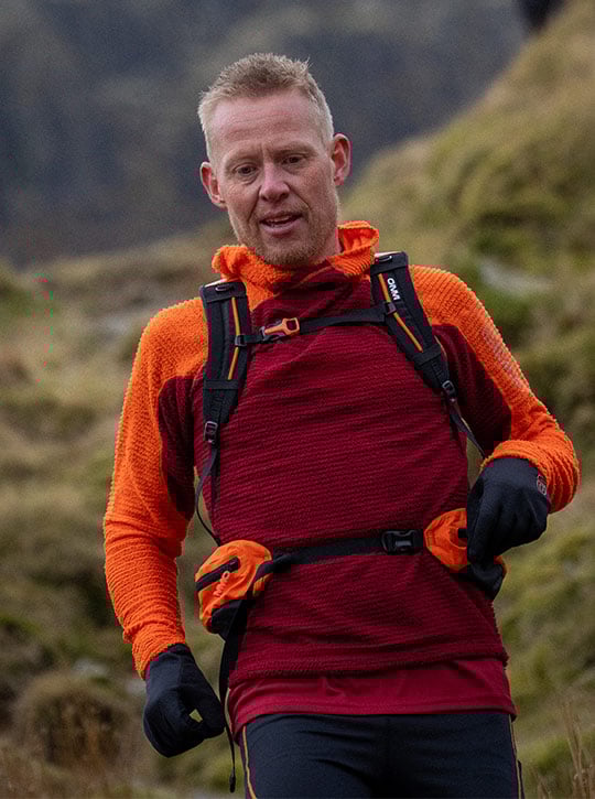 Man in orange and red hoodie runs down hill with a backpack in a mountainous area
