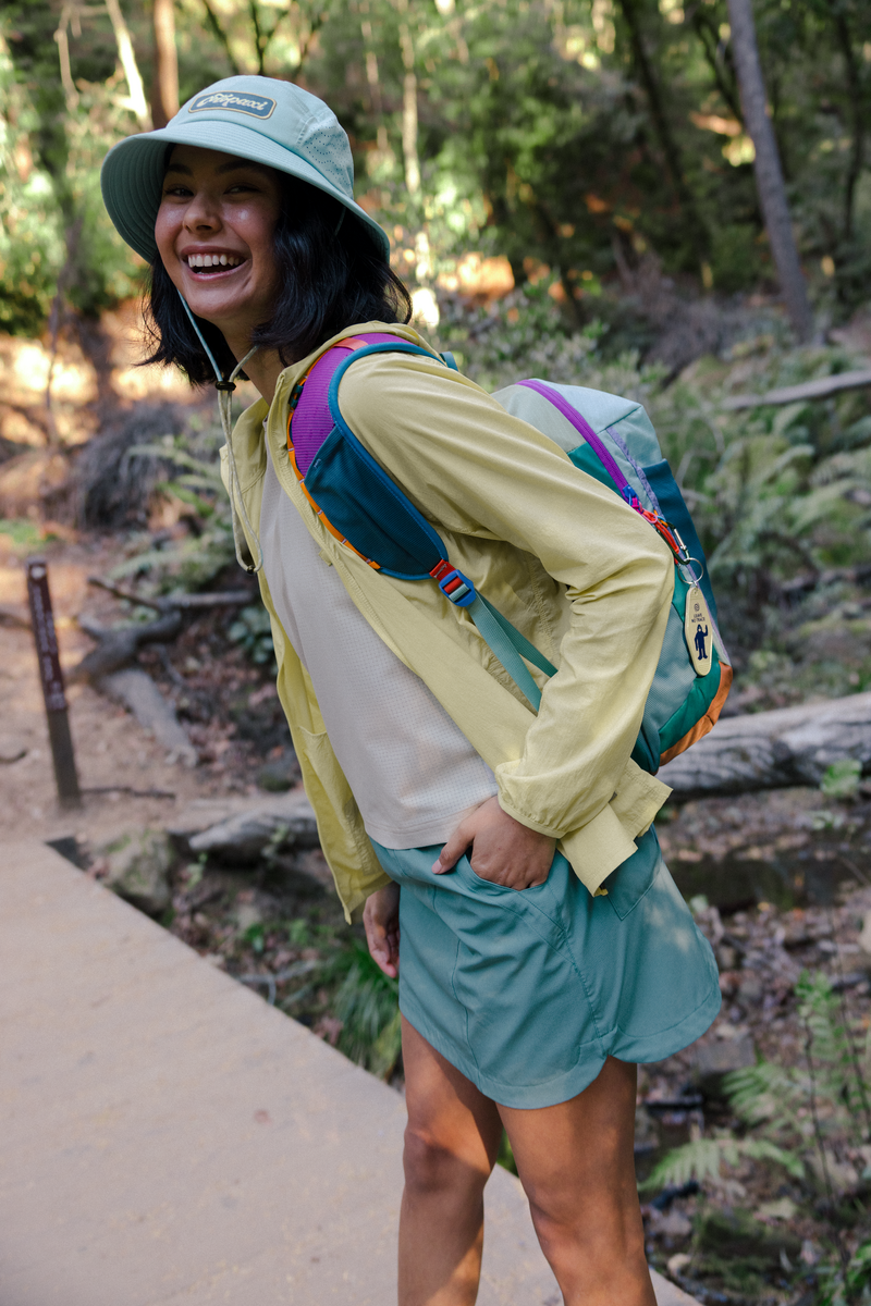 Person hiking in a forest wearing a yellow jacket and green shorts with a colorful backpack.