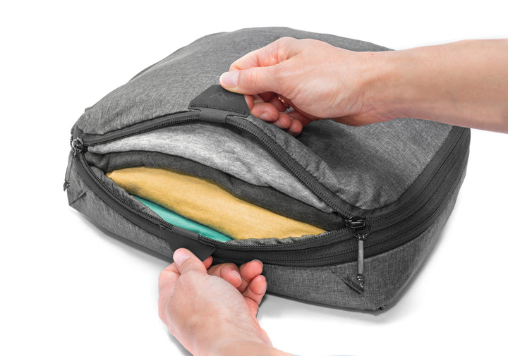 Gray travel bag with a person opening it to reveal clothes inside on a white background