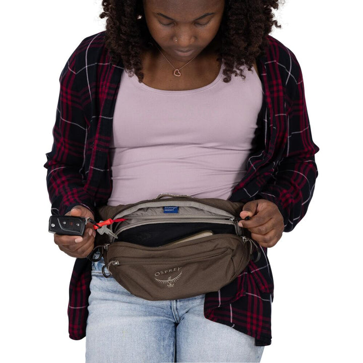 A woman wearing a brown Osprey waist bag, styled with a red and black plaid flannel shirt over a light pink top and light blue jeans. She is shown looking down into the open front pocket of the bag, which contains a black wallet and a red cord.