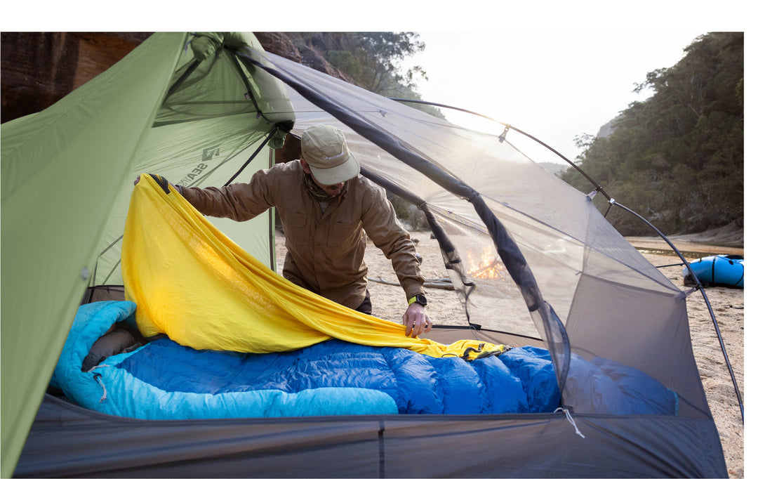 Person setting up a sleeping bag liner in a hiking tent. Driftwood fire and canoe in background