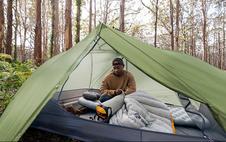 Person in a green tent in a forest setting, stuffing a sleeping bag into a compression sack