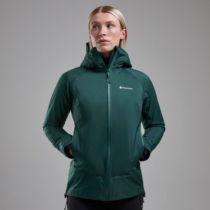 Woman wearing a dark green rain coat with the Montane logo, hands in high poockets