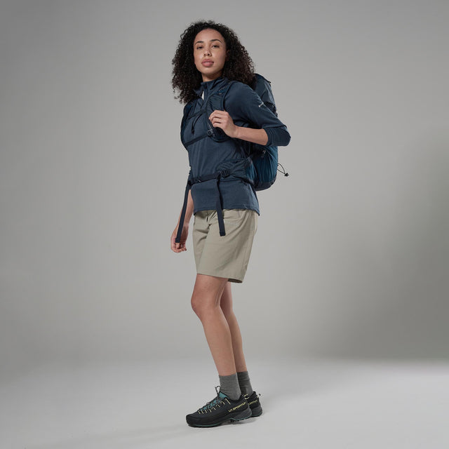 Person wearing a blue jacket and beige shorts with a backpack on a grey background