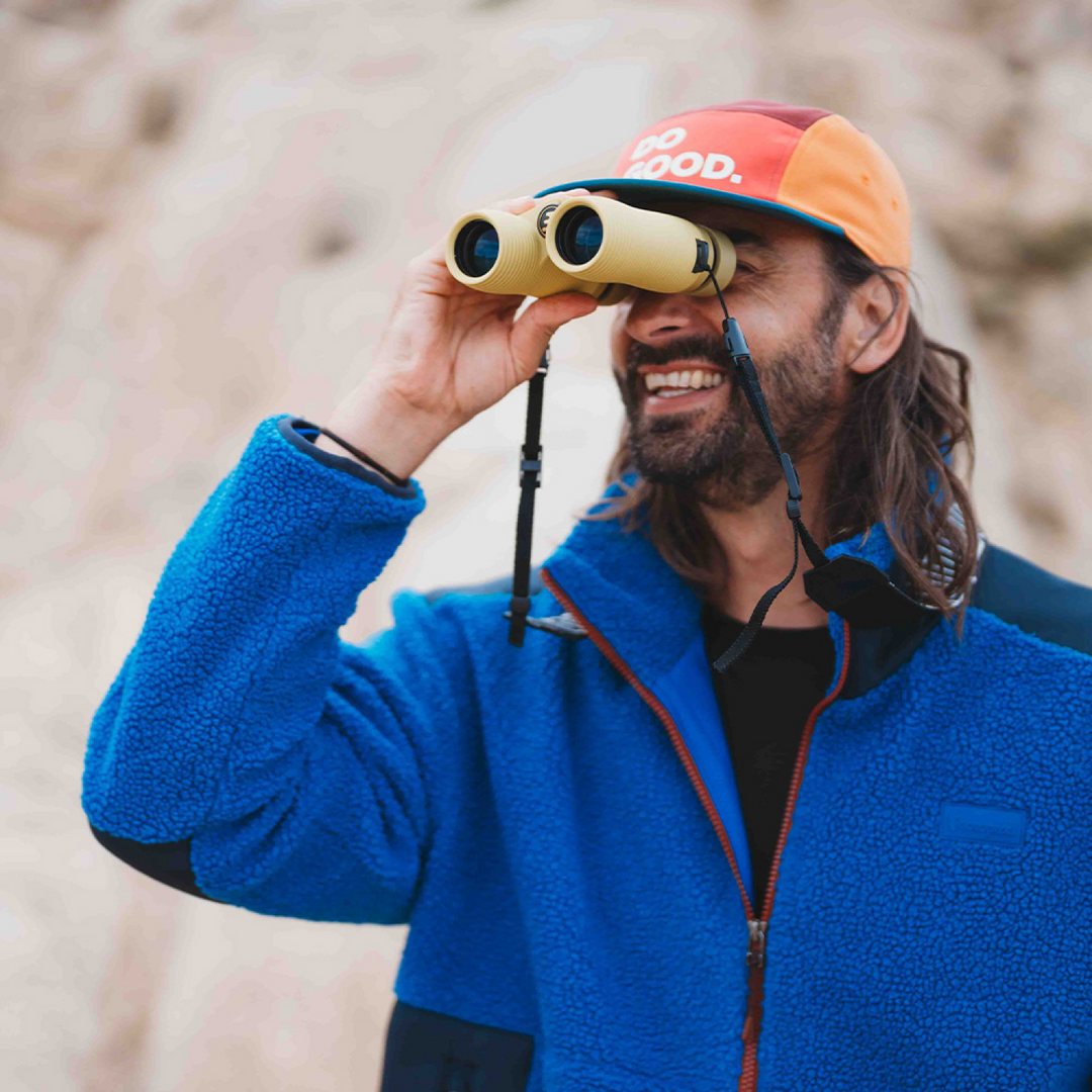 Man wearing a blue jacket in the outdoors, looking through a pair of off white binoculars.