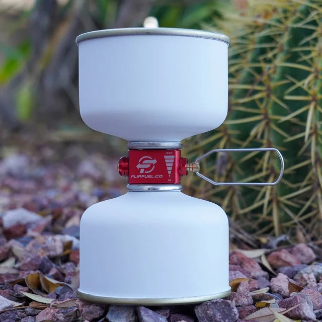 A white camping stove with a red fuel regulator and silver metal handle, set outdoors on a bed of small rocks with a blurred cactus in the background.