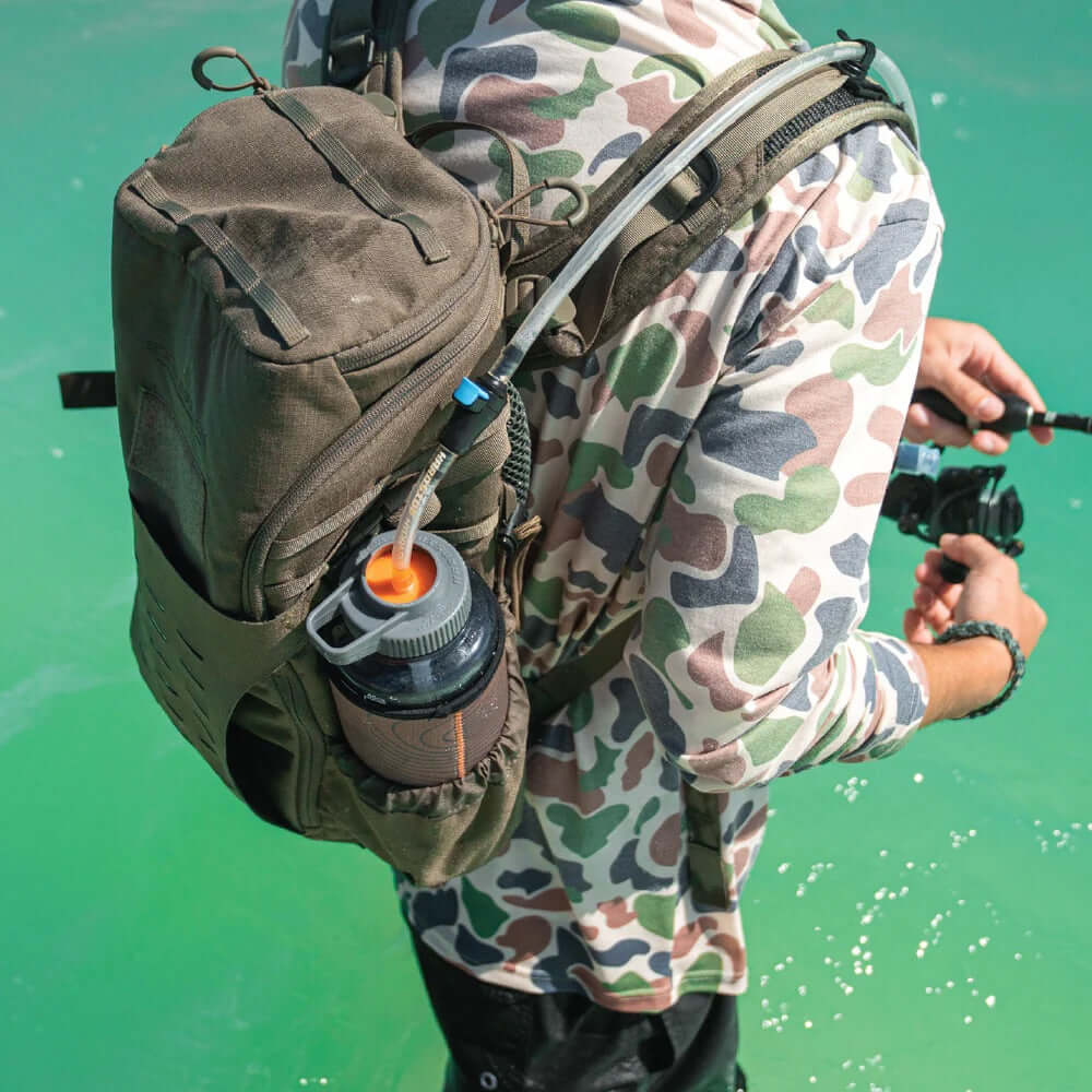 Person wearing a camouflage outfit with a backpack and water bottle with a hydration swig rig on a green background