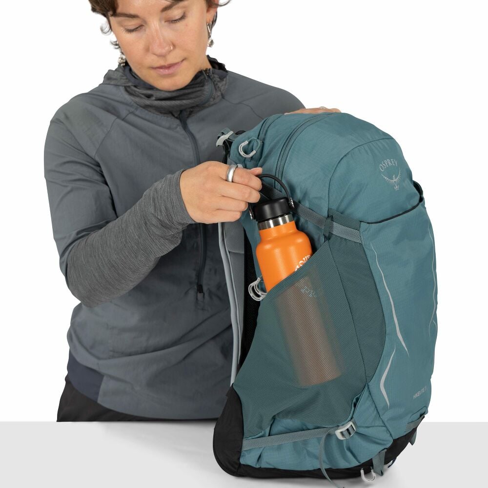 Person holding a teal backpack with an orange water bottle, wearing a gray jacket.