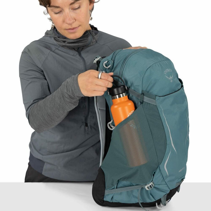 Person holding a teal backpack with an orange water bottle, wearing a gray jacket.