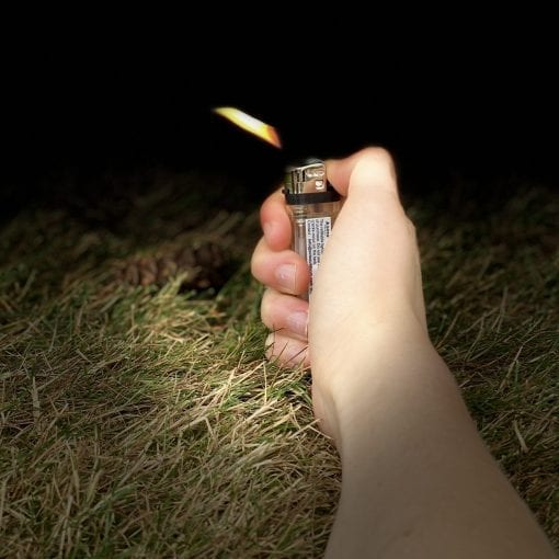 A hand holding a lighter outdoors at night, with a flame lit, over grassy ground in the foreground and a dark background.