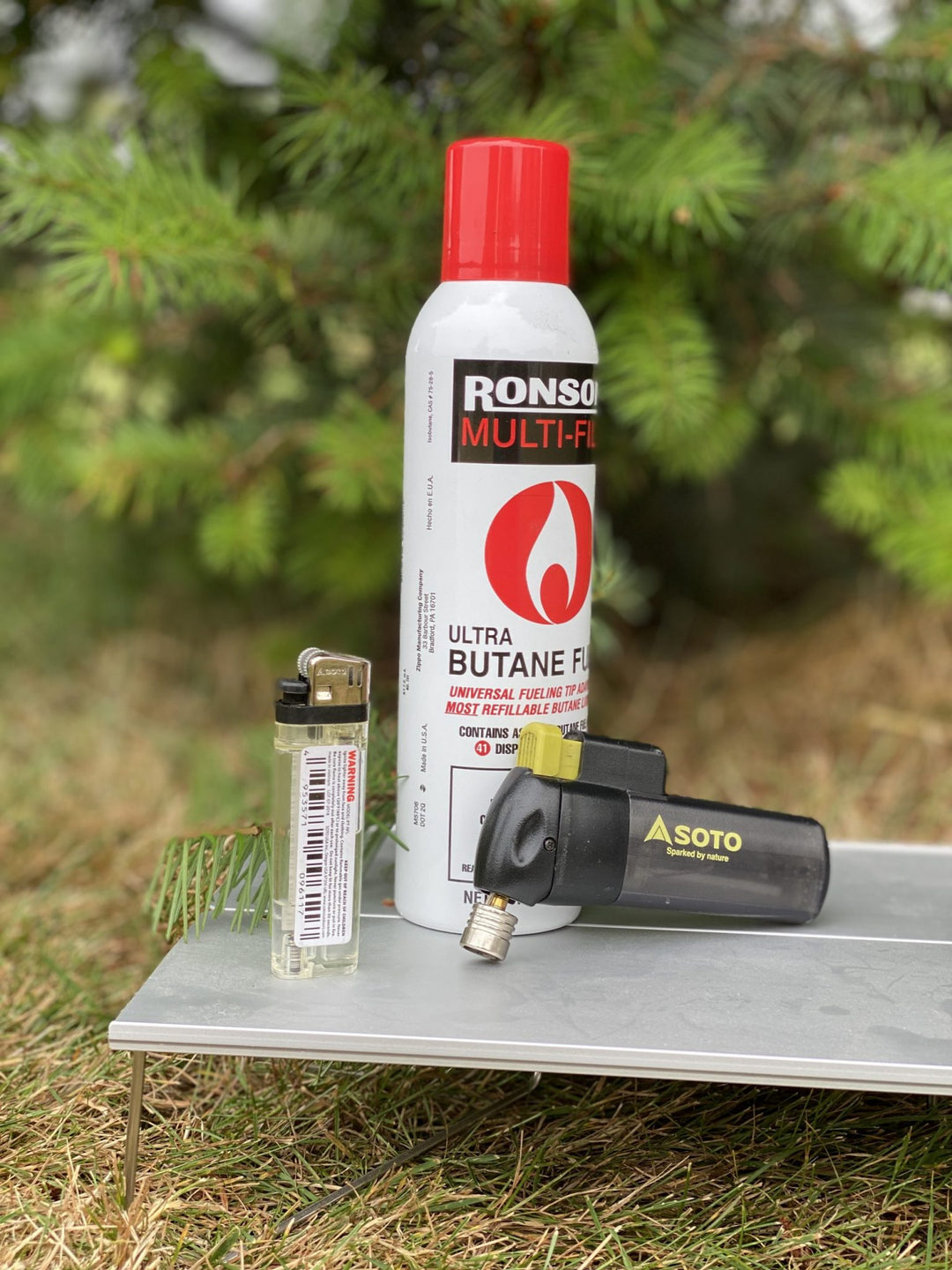 A can of Ronson Multi-Flame Ultra Butane Fuel stands on a metal table outdoors, with a small lighter and a black Soto torch lighter placed beside it against a blurred green foliage background.