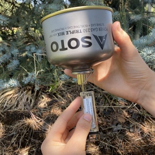 A person outdoors holds a silver SOTO Power Gas 230 triple mix camping stove burner, with the metal pot and fuel canister connected and a lighter held below the base. Pine trees and dry ground are visible in the background.