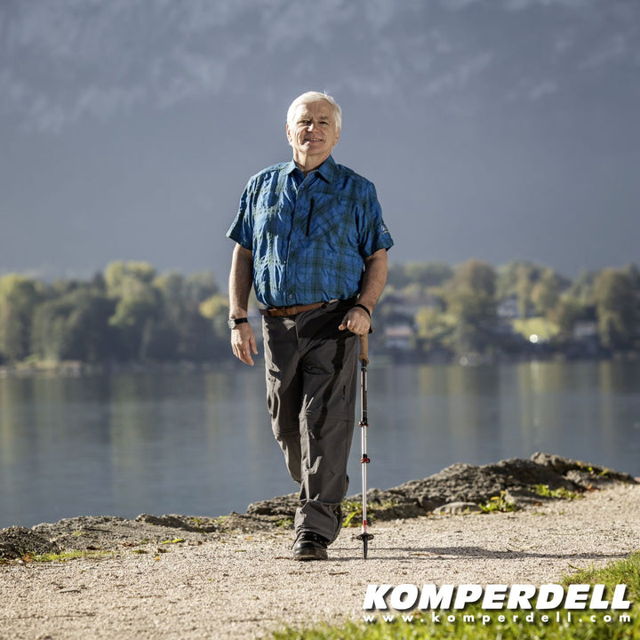 Elderly man walking along a gravel path beside a lake with a walking stick.
