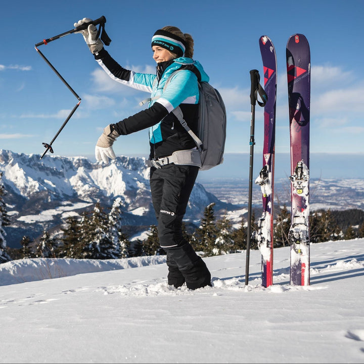 A woman skier stands on snowy mountain ground with ski poles and skis beside her. She wears a turquoise, black, and white ski jacket, black ski pants, a gray backpack, and a teal and black headband.