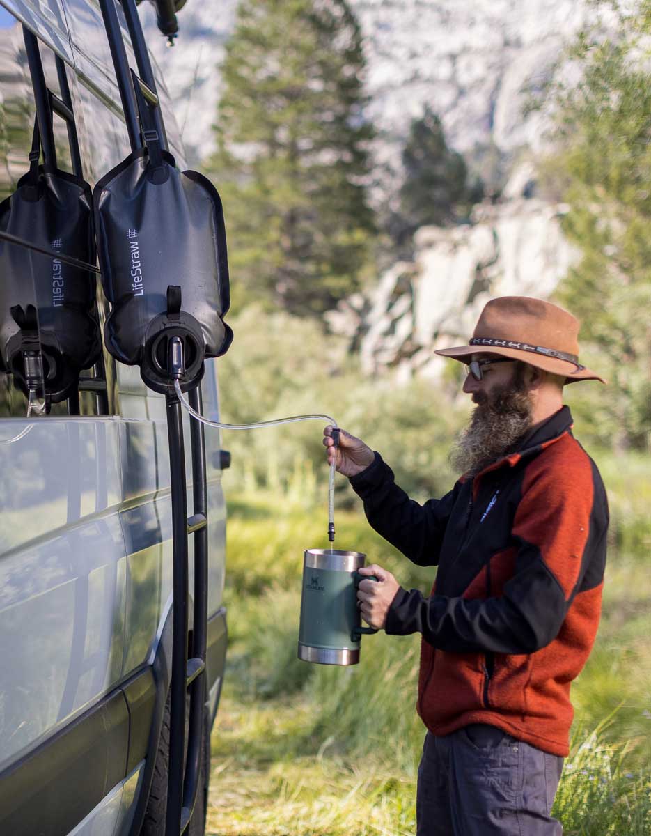 Man with a beard and hat holding a thermos near a van in a natural setting