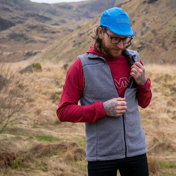 Man wearing a red jacket, gray vest, and blue cap in a mountainous landscape