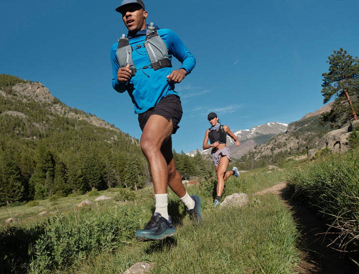 Two trail runners on a mountain path, with the foreground runner wearing a bright blue long-sleeve performance shirt, black shorts, and a hydration pack.