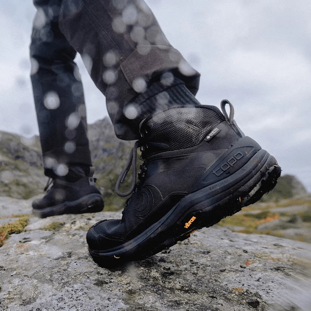 A close-up of a person wearing a black high-top waterproof hiking boot on a rocky outdoor trail, with raindrops visible on the lens and the brand name Vibram on the outsole.
