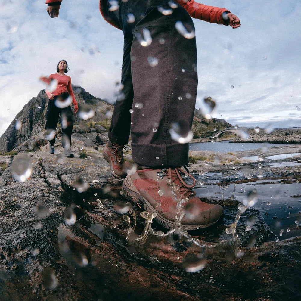 A low-angle outdoor shot of a person splashing through water on a rocky shoreline, with another person in the background near a mountain. The foreground person wears dark brown pants and reddish-brown hiking boots, with water droplets suspended in the air.