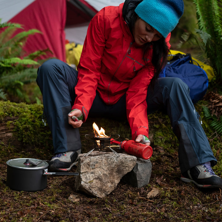 Person in red jacket and blue beanie preparing a campfire with a pot nearby, in a forest setting.