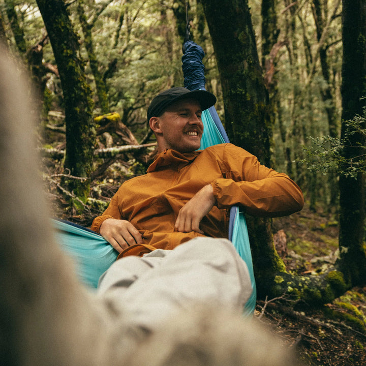 Man sitting in a blue hammock in a forest wearing an orange jacket. 