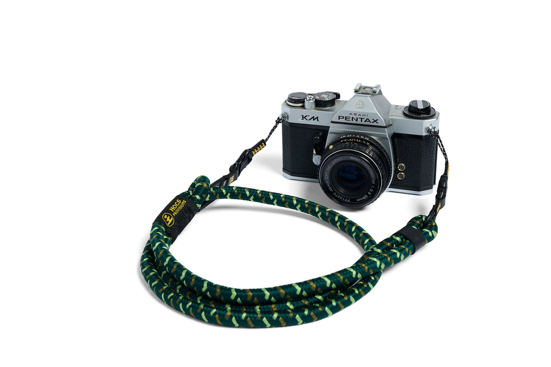 A green and black camouflage rope camera strap with yellow branding, shown draped around a silver and black Pentax K-M model camera on a white background.