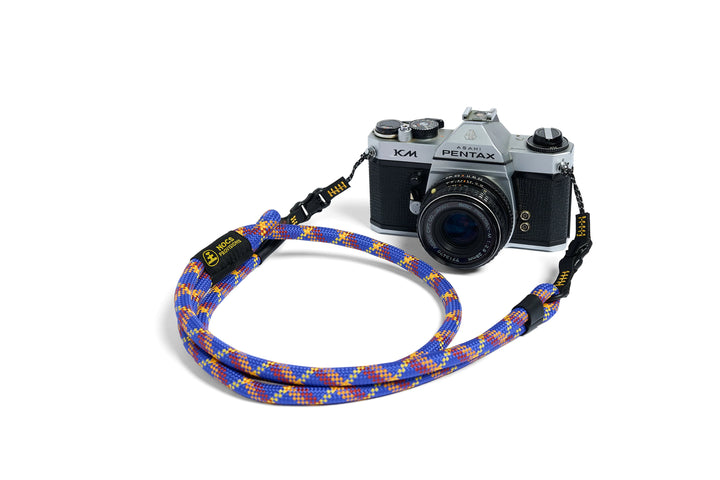 A multicolor braided camera strap with yellow text, shown carrying a silver and black Pentax K-Max camera on a clean white background.