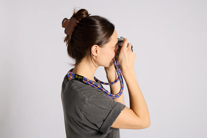 A woman in profile wearing a gray t-shirt and holding a camera, featuring a blue and black patterned camera strap with a black leather section around her neck.