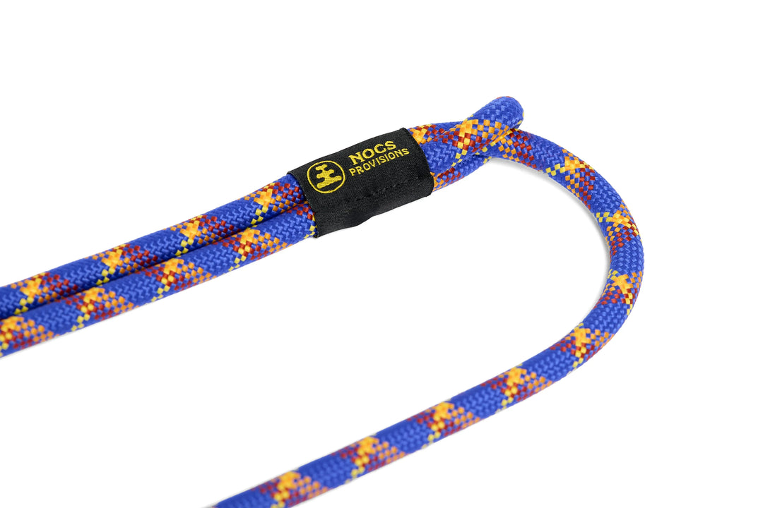 A close-up of a blue rope camera strap with yellow and red flecks, featuring a black Nocs Provisions label, shot against a solid white background.