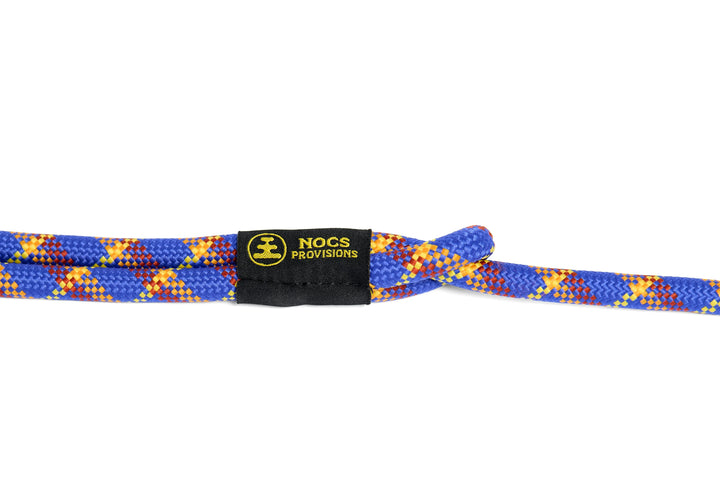 A close-up of a blue, yellow, and red patterned rope camera strap with a black NOCS PROVISIONS tag in the center, set against a plain white background.