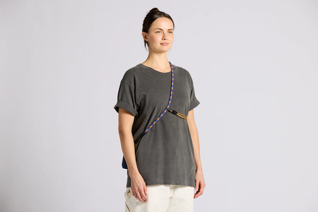 A woman modeling a dark gray oversized t-shirt with a colorful braided strap detail across the chest, paired with off-white pants, against a light gray studio background.