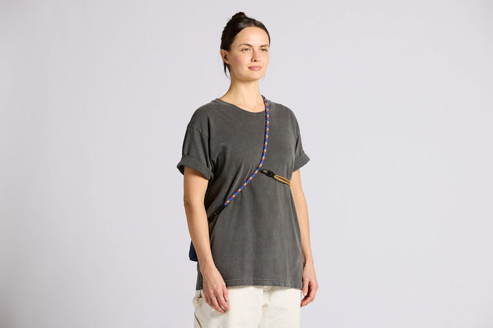 A woman modeling a dark gray oversized t-shirt with a colorful braided strap detail across the chest, paired with off-white pants, against a light gray studio background.