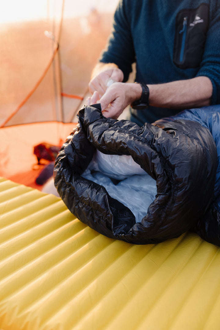 A close-up lifestyle shot of a person packing a black insulated sleeping bag onto a bright yellow camping mattress. The person is wearing a dark blue long-sleeve shirt, and the background shows a softly blurred campsite setting.