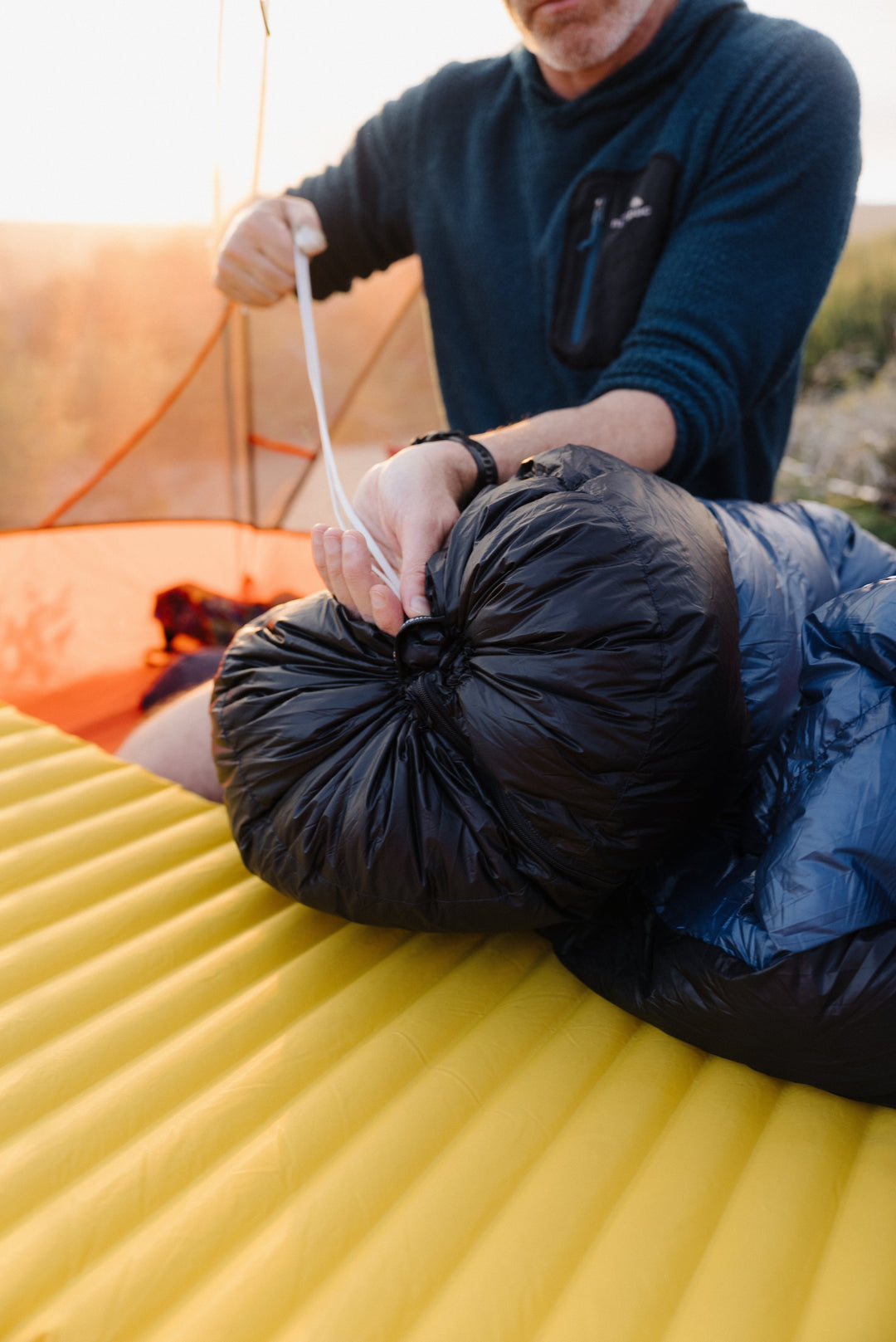 A man outdoors at sunset packs a black sleeping bag into a yellow sleeping pad, with an orange tent visible in the background.