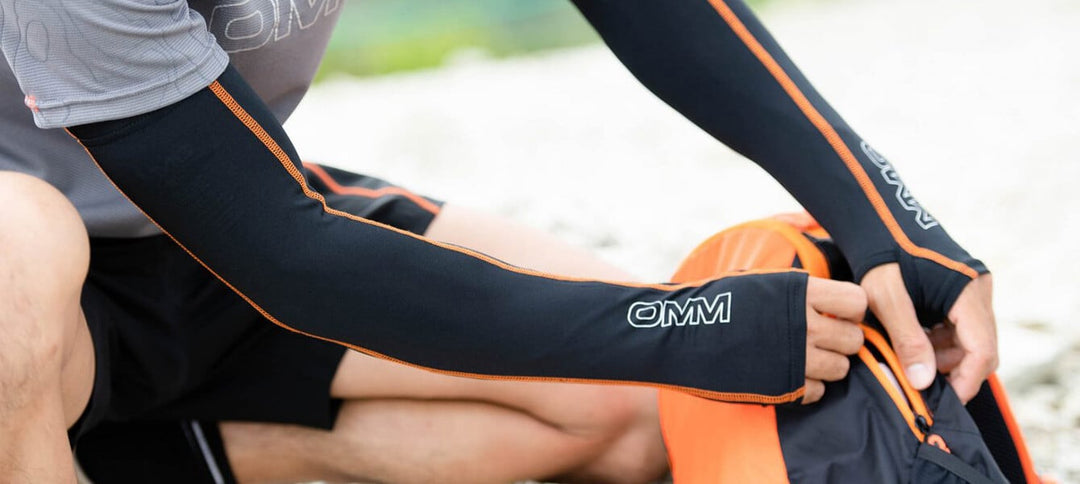 Person wearing black and orange arm warmes with 'OMM' branding, zipping closed a backpack in the outdoors