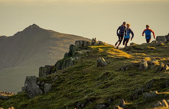 Three people running on a mountain trail with a scenic view of mountains and sky.